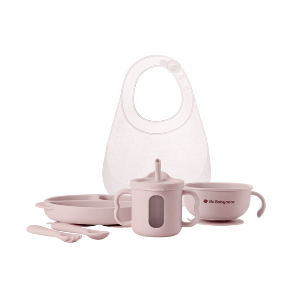White background image of a pink baby tableware set, including a bib, plate, fork and spoon, cup, and bowl.