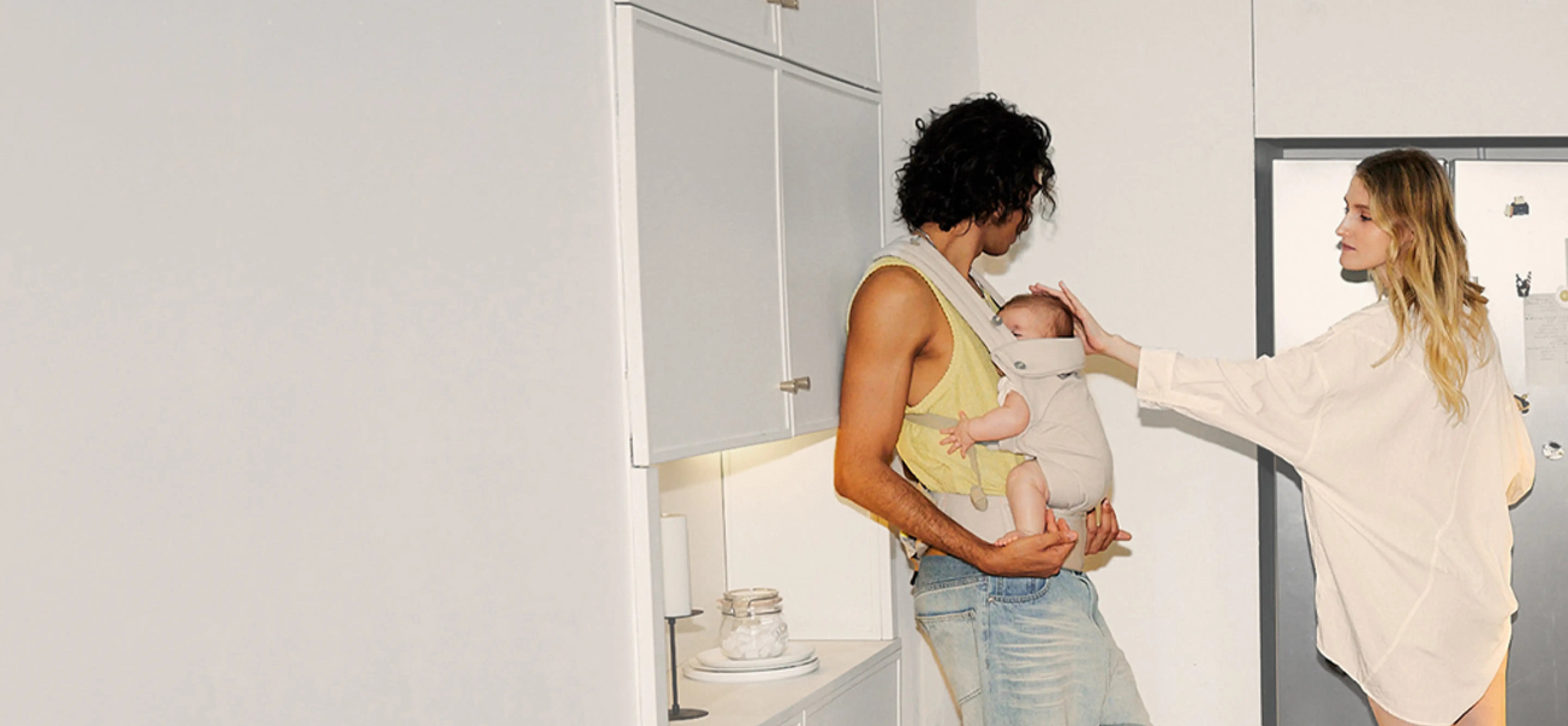 Father wearing the Bc Babycare CocoonGo Baby Carrier with a newborn while another parent stands nearby at home.