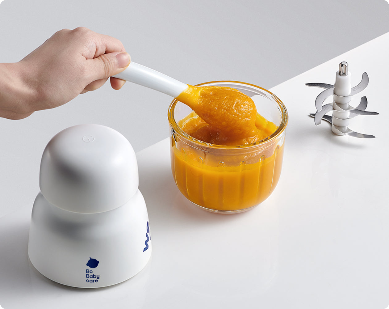 Smooth homemade orange baby puree in the glass bowl beside the BC Babycare Baby Food Maker and blade assembly.